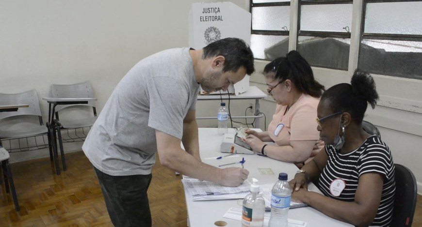 Eleitor preenche caderno de votação diante de mesa receptora de votos com duas mesárias