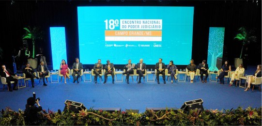 Foto mostra diversas pessoas sentadas em cadeiras sobre um palco